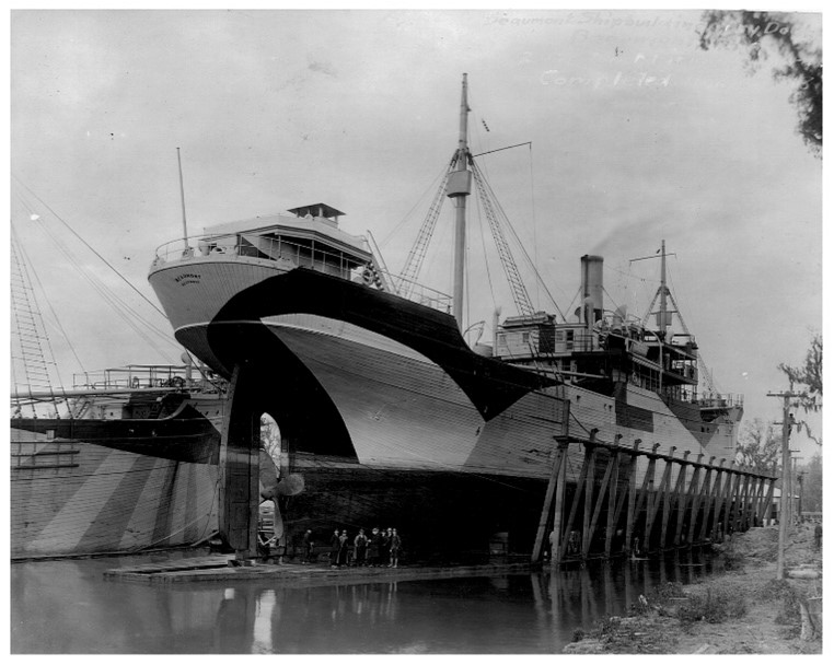 A wooden steamship is in a dry dock preparing to be launched at Beaumont in 1918. The black and white photographs shows the stern of the completed vessel with a second steamship in the river next to it. Both ships are painted in what was called a “dazzle” pattern to help camouflage the vessel at sea and make it hard to detect and sink by submarine.