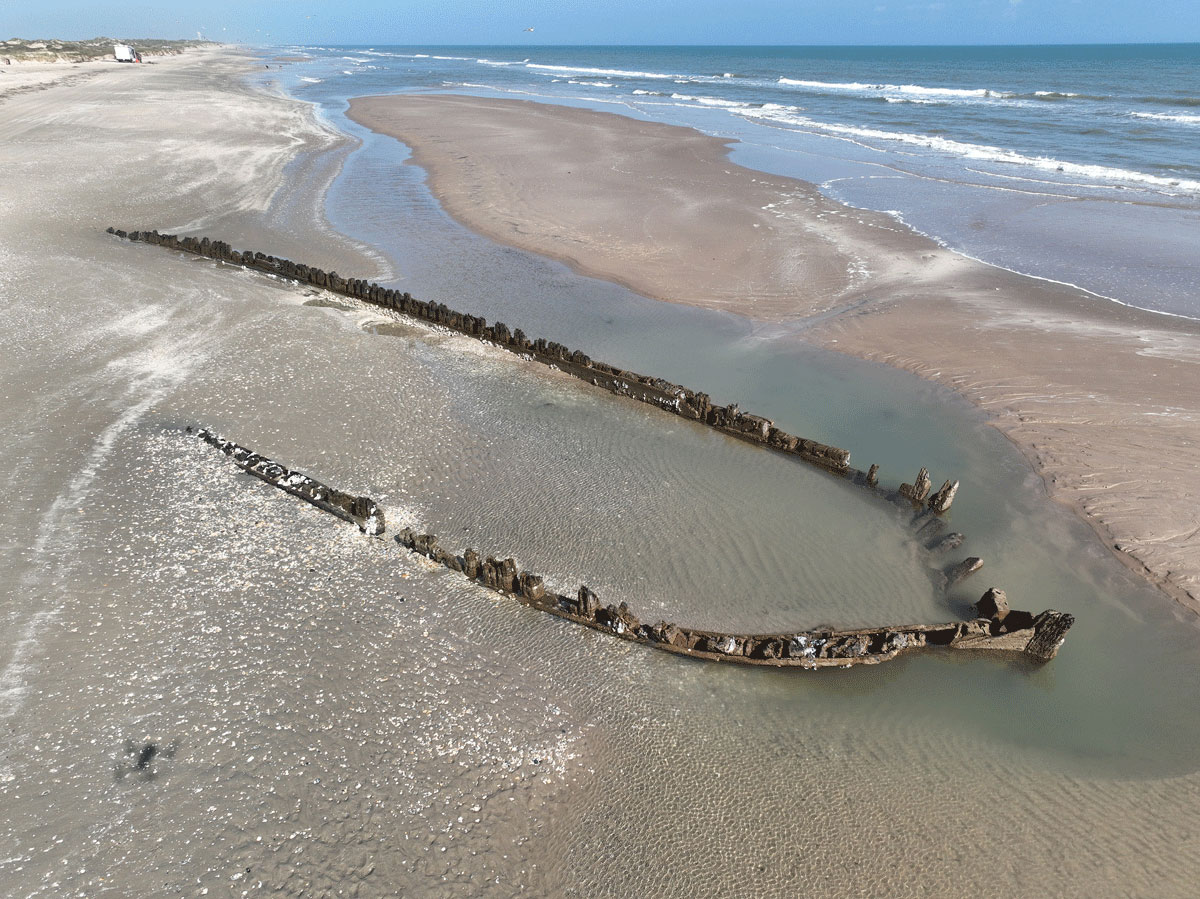 The lower portion of a wooden ship’s hull projects out of the sand on Boca Chica Beach, Texas in December 2022. The drone image shows the shipwreck on the beach, as seen from the air facing north, with the surf from the Gulf of Mexico approaching the shipwreck from the east.