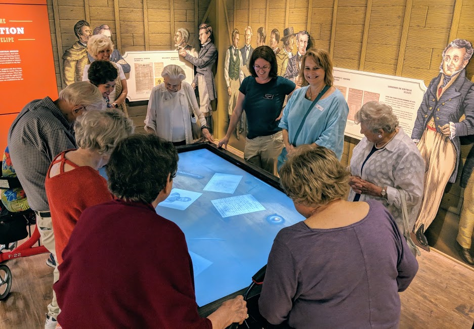 Photo displaying a group tour gathered around an interactive display in the center of the room