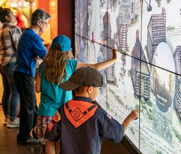 Three visitors on left side interacting with large, touch-screen wall of an illistration of the town of San Felipe de Austin as it may have looked 200 years ago