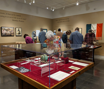 Collection of visitors facing away from camera looking at exhibit items. At forefront of image is a large case with plexiglass covering a variety of exhibits on display