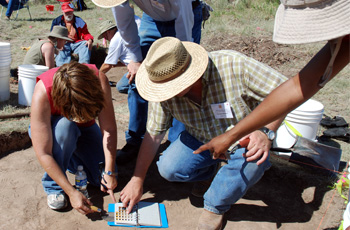 A man in a hat and others crouch down on the ground inspecting an archeological dig.