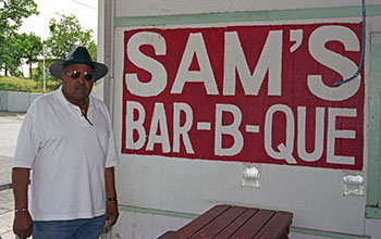 Picture of a man standing in front of a sign that reads "Sam's Bar-B-Que"